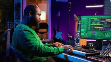 Male artist producing music on electronic piano keyboard in home studio. Sound engineer playing MIDI controller, recording and composing a soundtrack with acoustic tools and gear. Camera A. video