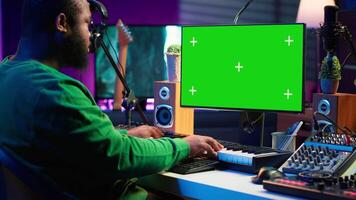 Producer composing music with electronic keyboard notes and green screen display on pc, post production in home studio. Musician artist plays midi controller next to mockup layout. Camera A. video