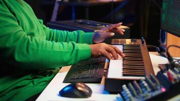 African american audio technician using piano synthesizer keys to add sound effects in post production, editing tunes with digital software and mixing console. Male artist in home studio. Camera A. video