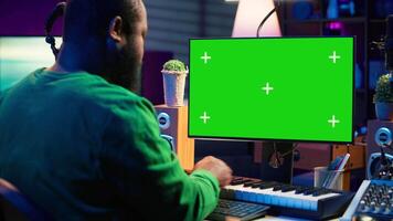 Musician operates on midi controller piano keys next to mockup screen, producing new songs with musical instruments and audio gear in home studio. Artist works with green screen. Camera A. video