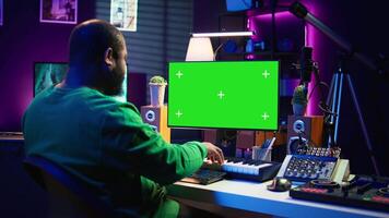 Producer composing music with electronic keyboard notes and green screen display on pc, post production in home studio. Musician artist plays midi controller next to mockup layout. Camera B. video
