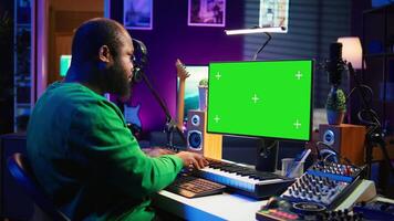 Musician operates on midi controller piano keys next to mockup screen, producing new songs with musical instruments and audio gear in home studio. Artist works with green screen. Camera B. video