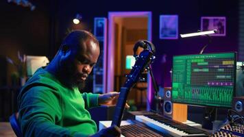 Producer composing new music on acoustic instrument in home studio, using microphone to sing chords. Musician songwriter works with mixing console and software, guitarist producing tunes. Camera B. video