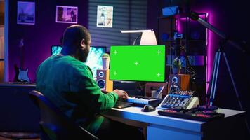 Songwriter operates on synthesizer piano keys next to isolated display, composing new music with instruments and stereo tools in home studio. Artist producer works with green screen. Camera B. video
