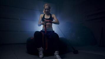 A boxer wraps bandages around his hands in preparation for a sparring match, in a dark room with a backlit room behind him video