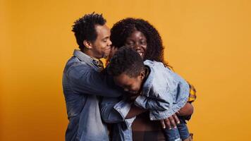 Joyful little child playing with a controller for games, posing with mother and father in studio. African american family with toddler standing against yellow background, kid gaming. Camera B. video