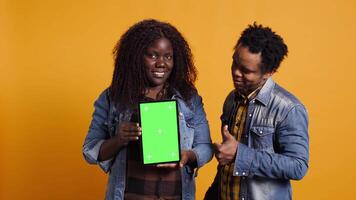 African american couple pointing at a tablet with isolated green screen display, smiling at each other against yellow background. Cheerful affectionate husband and wife posing. Camera B. video