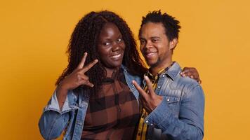 Cute life partners posing in studio and doing peace sign in studio, showing the victory symbol against yellow background. African american spouses doing kawaii gesture. Camera A. video