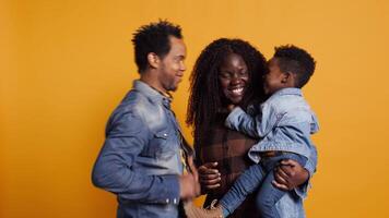 African american positive couple doing peek a boo with their young child in studio, smiling people hugging together. Happy affectionate mom and dad showing gratitude and respect. Camera B. video