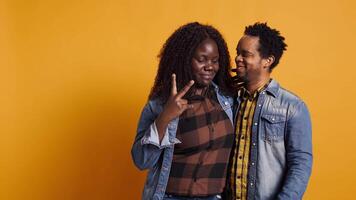 Cute life partners posing in studio and doing peace sign in studio, showing the victory symbol against yellow background. African american spouses doing kawaii gesture. Camera B. video