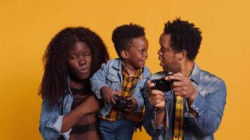 Happy small toddler playing around with a controller and sitting next to his parents, posing together in studio. African american cheerful family of three against yellow background. Camera A. video