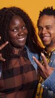 Vertical Cute life partners posing in studio and doing peace sign in studio, showing the victory symbol against yellow background. African american spouses doing kawaii gesture. Camera A. video