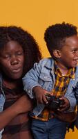 Vertical Happy small toddler playing around with a controller and sitting next to his parents, posing together in studio. African american cheerful family of three against yellow background. Camera A. video