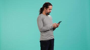 Happy man texting best friend using smartphone, reading messages. Relaxed person having fun while chatting with mate on mobile phone, isolated over blue studio background, camera A video