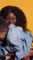 Vertical Joyful little child playing with a controller for games, posing with mother and father in studio. African american family with toddler standing against yellow background, kid gaming. Camera A. video