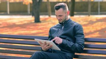 Student is Reading News or Watching on Tablet Computer in the Park in Autumn. Man in Black Leather Jacket is Sitting on a Bench Using Big White Tablet PC. Middle Close-Up Shot video