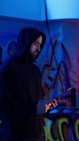 Vertical Lone wolf hacker in neon lit secret hideout trying to hide his online activity after stealing data from victims using computer virus, trying to avoid being detected, using tablet, camera B video