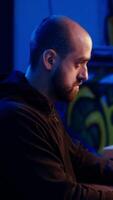 Vertical Hacker building spyware software designed to gather information from users computers without their knowledge. Man doing cybercriminal activities in rundown hideout, dolly out shot video