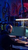 Vertical Hacker in underground warehouse writing lines of code on computer screen to make malware that get past security systems. Man in hidden HQ building scripts that can hack devices, dolly in shot video