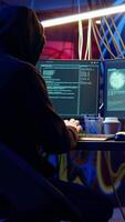 Vertical Hooded man in underground hideaway listening and breaking into computer systems to access valuable data. Evil hacker wearing headphones bypassing firewalls and login process, panning shot video
