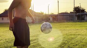 Young man juggling soccer ball on stadium at sunset. Professional footballer kicking ball at green field. Sportsman practicing tricks at meadow with sunlight at background. Freestyle football video