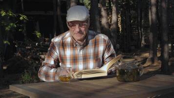 An elderly man sits at a wooden table, savoring a cup of tea while reading a book. Surrounded by trees, he enjoys a tranquil moment in nature, immersed in his literature. video