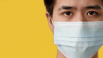 A young Asian guy displays a serious expression, wearing a surgical mask against a vibrant yellow backdrop, symbolizing health and safety in contemporary settings. video
