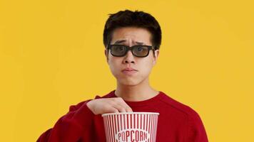 The young Asian man, wearing glasses and a red sweater, excitedly eats popcorn from a classic striped container, clearly immersed in a fun movie experience in a vibrant environment. video
