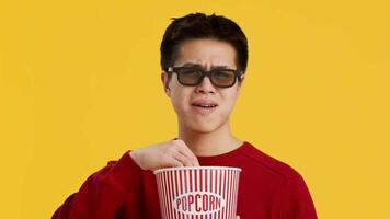 A young Asian man wearing glasses reaches into a striped popcorn bucket, looking intrigued while enjoying a movie. His colorful sweater contrasts with the vibrant yellow background. video