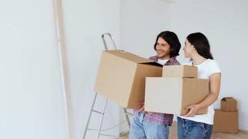 A couple carries cardboard boxes into their new home, preparing for a renovation. They are smiling and engaged, surrounded by a partially painted wall and a ladder. video