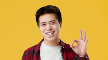 Asian young man is happily smiling while making an okay hand gesture. He stands in front of a vibrant yellow background, exuding positivity and confidence. video
