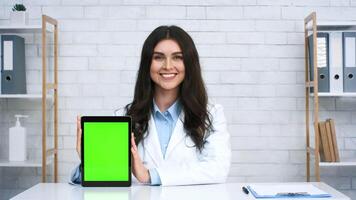 A smiling woman doctor in a white coat showcases a tablet with a blank green screen. The setting is a bright office with shelves and a clipboard visible, creating a welcoming atmosphere. video