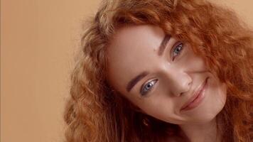 A cheerful young woman with bright blue eyes and vibrant curly red hair smiles warmly, creating an inviting atmosphere in a softly lit setting with a neutral background. video