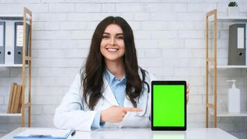 A healthcare professional in a white coat sits at a desk in a clinic, smiling and pointing at a digital tablet displaying a green screen, ready for information or graphics. video