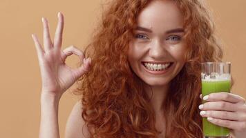 A cheerful woman with vibrant red curls holds a glass of green smoothie, flashing an enthusiastic okay sign. Her bright smile and lively expression convey joy and satisfaction. video