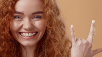 A lively young woman with curly red hair smiles broadly while making a hand gesture associated with excitement. The background is a warm, neutral color, enhancing her joyful expression. video