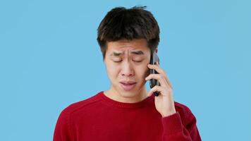Asian young man wearing a red sweater converses on his cellphone, looking worried. The background is a solid blue, emphasizing his expression and the seriousness of the conversation. video