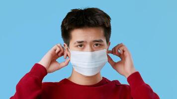 Asian young man is adjusting a medical mask on his face, preparing to wear it. He is focused and appears to be in a well-lit space with a solid blue backdrop, highlighting his actions. video