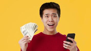 Asian young man in a red sweater displays a shocked expression as he holds a stack of money in one hand and looks at his phone with astonishment, set against a vibrant yellow background. video