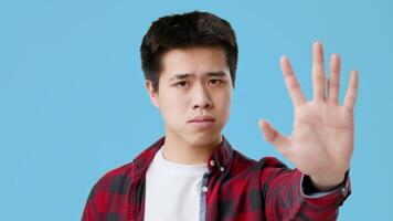 Asian young man stands against a blue backdrop, displaying a serious demeanor while raising his hand as a gesture of stopping or caution. He wears a casual shirt and looks directly at the viewer. video
