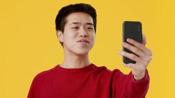 Asian young man dressed in a red sweater happily waves at his smartphone while smiling against a vibrant yellow background, showcasing a cheerful moment of connection or communication. video