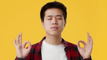 Asian young man sits cross-legged with a calm expression, practicing meditation. He is dressed casually in a checkered shirt and white tee, immersed in a serene moment of mindfulness. video