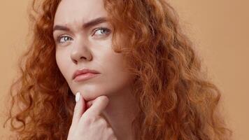 A woman with striking curly red hair contemplates while resting her chin on her hand. Her expression suggests deep thought or skepticism, set against a warm background. video
