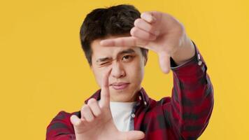 Asian young man is creating a framing gesture with his hands while standing in front of a vibrant yellow backdrop. He appears focused and engaged, ready to express creativity. video