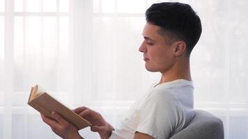 A young man sits comfortably in a light-filled room, immersed in a book. The sunlight streaming through the window creates a warm and serene atmosphere, perfect for reading. video