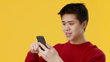 Asian young man is focused on his smartphone, intently reading or interacting with an app. The bright yellow background adds an energetic feel to the indoor setting. video