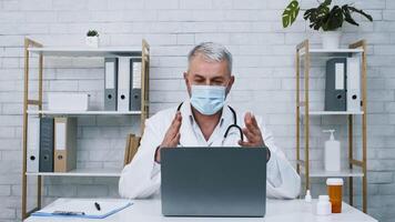 A healthcare professional in a lab coat and mask is engaged in a virtual consultation. He is seated at a desk with a laptop, discussing health matters and documenting notes in a clinical setting. video
