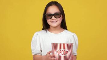 A cheerful Asian girl holds a striped popcorn bucket and wears 3D glasses, set against a bright yellow backdrop. video
