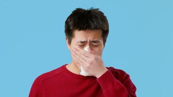 Asian young man in a red sweater is indoors, visibly uncomfortable while holding a tissue to his face. He appears to be dealing with allergy symptoms in a light blue setting. video