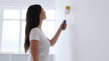 A woman is focused on painting a wall bright white using a roller. Sunlight streams through large windows, creating a cheerful and inviting atmosphere in the room. video